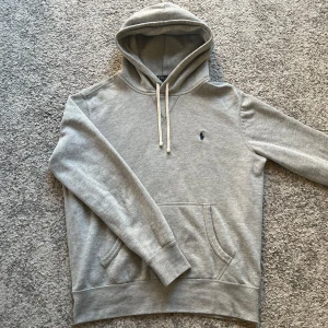 Ralph Lauren hoodie  - Säljer en klassisk grå hoodie från Polo Ralph Lauren med broderad marinblå logga på bröstet. Tröjan har huva med dragsko och en stor magficka. Perfekt för en avslappnad stil.