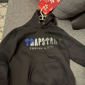  Trapstar hoddie,strl M - Säljer en svart hoodie från Trapstar med texten 'TRAPSTAR IT'S A SECRET' i blått och grått på framsidan. Hoodien har en klassisk design med dragsko i huvan och ribbade muddar vid ärmslut och nederkant.