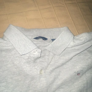 Ljusgrå pikétröja från GANT - Säljer en klassisk ljusgrå pikétröja från GANT med broderad logga på bröstet. Tröjan har korta ärmar, krage och knäppning framtill. Perfekt för en stilren och avslappnad look.