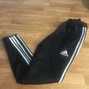 Svarta Adidas mjukisbyxor med vita ränder - Säljer ett par svarta Adidas mjukisbyxor med klassiska vita ränder längs sidorna och dragkedjefickor. Byxorna har resår i midjan och är perfekta för en sportig look.