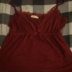 Vinröd linnetopp med spets från Nelly - Säljer en vinröd linnetopp från Nelly med tunna axelband och fina spetsdetaljer längs urringningen och nederkanten. Toppen har en liten rosett framtill och är perfekt för en trendig och feminin look. Har ni fler frågor är det bara att skriva💕