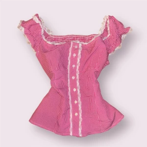 Rosa rutig blus med spetsdetaljer - Supersöt rosa och vit rutig blus med volangärmar och spets längs kanterna. Knappar framtill och figurnära passform. Perfekt för dig som vill ha en romantisk och lekfull stil.