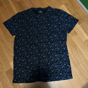 Svart t-shirt med blommigt mönster från Dressmann - Säljer en svart t-shirt från Dressmann i storlek M. T-shirten har ett diskret blommigt mönster i mörkblått och är tillverkad i mjukt material. Perfekt för dig som vill ha en stilren men ändå unik look.