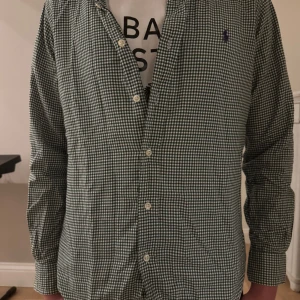 Rutig grön Ralph lauren skjorta  - Klassisk långärmad skjorta från Ralph Lauren i grönt och vitt rutigt mönster. Skjortan har knappar framtill och en liten broderad logga på bröstet. Perfekt för en stilren och avslappnad look. Inprincip helt ny. Mitt pris är 399kr. 