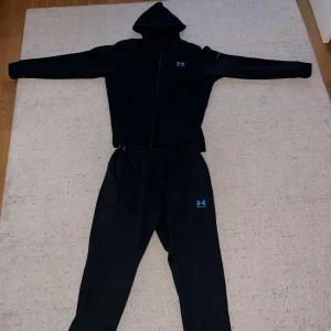 Svart hoodie från Under Armour - Säljer en svart hoodie från Under Armour med dragkedja och huva. Tröjan har en diskret blå logga på bröstet och är perfekt för träning eller chill. Klassisk passform och långa ärmar.