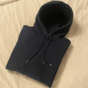 Mörkblå hoodie från Tommy Hilfiger - Säljer en stilren mörkblå hoodie från Tommy Hilfiger med liten broderad logga på bröstet. Tröjan har huva med dragsko och är perfekt för en avslappnad look.
