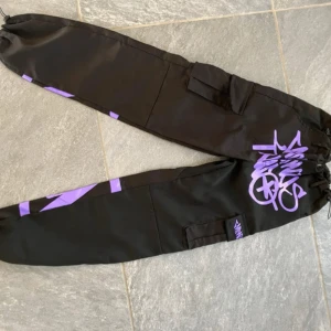 Svarta Minus Two cargpants med lila tryck - 🏷️ Brand: Minus Two 📐 Size: M 🎨 Color: Black, Purple 🧵 Material: Polyester 👕 Fit: Regular 📏 Length: Regular ⭐ Condition: Like new, Worn a few times ⚠️ Defects: None