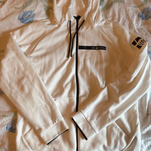 Vit hoodie från Sail Racing - Vit hoodie från Sail Racing med svart dragkedja och snörning i huvan. Jackan har två fickor framtill och en logotyp på vänster ärm samt bröstet. Perfekt för en sportig och avslappnad stil.