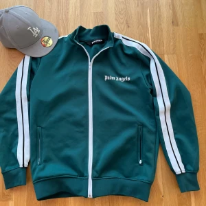 Palm Angels zip hoodie - Säljer en grön track jacket från Palm Angels med vita ränder längs ärmarna och vit dragkedja. Jackan har ståkrage och två fickor framtill. Klassisk logga på bröstet. Perfekt för en sportig och trendig look. Som ny inget trasigt bra passform hör av dig om du intresserad 