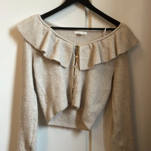 Beige stickad kofta med volang - Superfin beige stickad kofta med bred volang runt axlarna och knappar framtill. Koftan är croppad och har långärmade ärmar, perfekt för dig som vill ha en söt och trendig look.