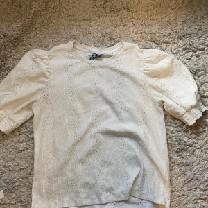 Vit blus med puffärm från Pieces - Mycket bra skick!! Supersöt vit blus från Pieces med korta puffärmar och vågigt broderat hålmönster. Blusen har rund halsringning och resår vid ärmslut för extra volym. Perfekt för dig som gillar romantiska detaljer och en ljus, fräsch stil.