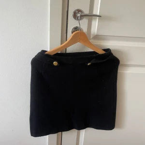 Svart stickad kjol från Bondelid - Svart stickad kortkjol från Bondelid med snygga guldknappar i midjan. Klassisk och enkel design som är lätt att matcha. Perfekt för dig som gillar stilrena plagg med en liten detalj som sticker ut.