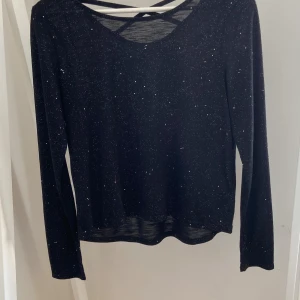 Svart glittrig långärmad topp från H&M - Svart långärmad topp med glittriga detaljer och snyggt korsade band i ryggen. Tunt och lätt material som ger en cool look. Perfekt för dig som vill sticka ut lite extra med en touch av glitter.