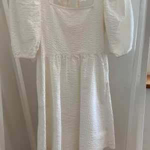 Vit kortklänning Holly & Whyte  - Superfin vit kortklänning från Holly & Whyte med puffärmar och snygg knytning i ryggen. Klänningen har en strukturerad yta och fyrkantig ringning, perfekt för sommarens alla tillfällen. Söt och trendig modell med volym i ärmarna.