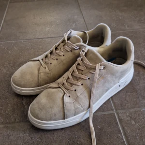 Beige mockasneakers med vit sula - Stilrena sneakers i beige mocka med vita sulor och matchande beige snörning. Skorna har en klassisk rund tå och platt sula, perfekt för en clean och avslappnad look. Passar dig som gillar neutrala färger och enkel design.