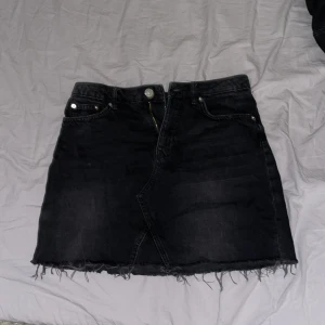 Svart jeanskjol med fransig kant - Svart kort jeanskjol med rå fransig kant nertill och klassiska fickor både fram och bak. Kjolen har dragkedja och knapp framtill samt bälteshällor. Perfekt för en avslappnad sommarkväll