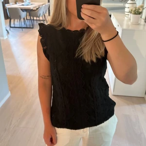 Svart spetsblus med volangärm - Supersnygg svart blus i spets med vågigt mönster och volangdetaljer vid axlarna. Ärmlös modell med rundad halsringning och feminina spetskanter. Perfekt att styla med både jeans och kjol för en trendig look. Aldrig använd utan prislapp 