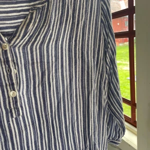 Randig blåvit henley i linne - Snygg henley-skjorta med blå och vita vertikala ränder. Skjortan har trekvartsärm, knappar framtill och är tillverkad i svalt linne. 