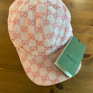 Rosa keps från Gucci - Snygg rosa keps från Gucci med ikoniskt GG-mönster i en ljusrosa och mörkare rosa ton. Justerbart band bak i rosa skinn med guldfärgad spänne. Kepsen har märkets logga på sidan. Perfekt för dig som vill sticka ut med lyxig streetstyle. Helt ny icke andvänd! 