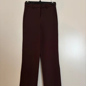 Bruna kostymbyxor från Vero Moda - Snygga bruna kostymbyxor från Vero Moda med raka ben och midjehällor. Byxorna har klassisk design med diskreta fickor i sidorna och stängs med dold dragkedja fram. Perfekta för en clean och stilren look.