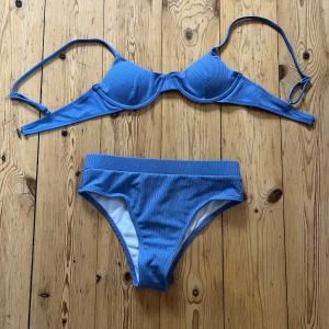 Blå ribbad bikini i storlek S - Snygg blå bikini med ribbad struktur och bygel-bh. Överdelen har justerbara axelband och spänne i ryggen. Underdelen är klassisk med bredare midjeband. Tillverkad i stretchig polyesterblandning. Säljer då den är för liten för mig. Den är lite trasig vid spännet men funkar ändå utmärkt.