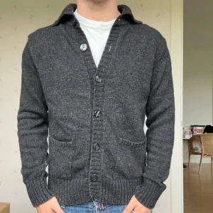 Mörkgrå stickad kofta från Oscar Jacobsson - Snygg exklusiv stickad kofta från Oscar Jacobsson med grov ribbad krage och knäppning framtill. Två praktiska fickor och breda muddar ger en chill och klassisk look. Perfekt för lager-på-lager under kyliga dagar. Hör av dig vid frågor och funderingar! 