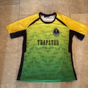 Trapstar grön/gul fotbollströja S - Säljer en snygg fotbollströja från Trapstar i storlek S. Tröjan har grön framsida med grafiskt mönster, gula ärmar och svarta detaljer. Tryck med Trapstar-logga och nummer 23 på ryggen. Tillverkad i lätt och ventilerande polyester, perfekt för match eller träning.