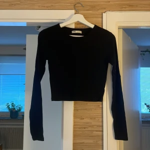 Svart croppad långärmad topp Pull&Bear - En svart, croppad långärmad topp från Pull&Bear. Enkel och stilren design med rund halsringning och tight passform. Perfekt att matcha med höga jeans eller kjol för en clean look.