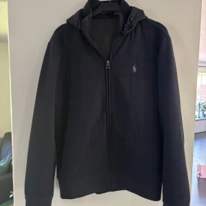 Mörkblå vindjacka från Polo Ralph Lauren - Nypris 3900 kr. Mörkblå vindjacka från Polo Ralph Lauren med huva och dragkedja framtill. Jackan har diskret logga på bröstet och är tillverkad i ett mjukt material med slät framsida och stickade ärmar. Perfekt för dig som gillar stilren och sportig look.