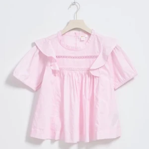 Ljusrosa blus med volang från Gina Tricot - Supersöt ljusrosa blus från Gina Tricot med korta puffärmar, volangdetaljer vid axlarna och broderad spets över bröstet. Blusen har rund halsringning med liten volangkant och knäppning bak i nacken🩷