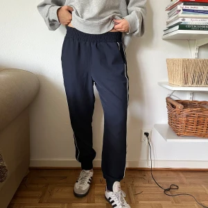 Mörkblå träningsbyxor med vita ränder - Snygga blåa träningsbyxor med vita ränder längs sidorna. De har elastisk midja, raka ben och muddar vid anklarna. Perfekta för en chill och sportig look. Materialet känns mjukt och bekvämt, och de har praktiska fickor på sidorna.