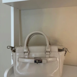 Beige axelväska med låsdetalj - Snygg beige axelväska med både handtag och justerbar axelrem. Väskan har silverfärgade metalldetaljer, dragkedja upptill och en dekorativ låsdetalj framtill. Perfekt storlek för det viktigaste och har innerfack för småsaker.🫶🏻💗