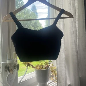 Svart korsett-topp med dragkedja - Snygg svart korsett-topp med breda axelband och cool dragkedja bak. Toppen har en croppad passform och detaljer med utskurna partier på ryggen som ger en edgy vibe. Perfekt att styla med höga jeans eller kjol för en trendig look.