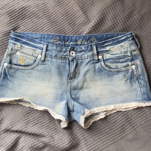 Lågmidjade Jeansshorts med spets - Sjukt söta och trendiga ljusblå lågmidjade jeansshorts med snygga detaljer och vit spetskant längst ner. Köpta på loppis och är i bra skick. I shortsen står det 27 och jag skulle säga att de passar XS/S Det är bara att skriva vid frågor!