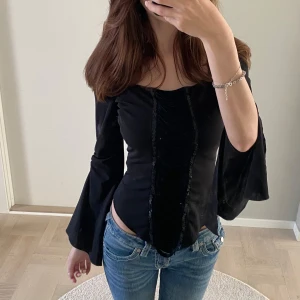 Svart offshoulder topp med utsvängda ärmar - Supersnygg svart topp med offshoulder och smala axelband. Utsvängda ärmar och coola fransdetaljer framtill. Figurnära passform och lätt croppad med spetsig nederkant.