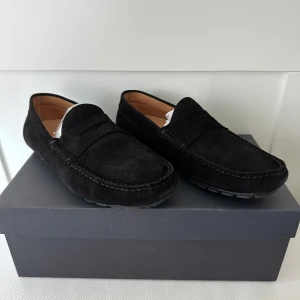 Svarta mockaloafers från Novita - Stilrena svarta loafers från Novita i mjuk mocka. Skorna har klassisk design med rund tå och diskret söm längs kanten. Perfekta för en clean och dressad look, med bekväm platt sula och snygg finish.