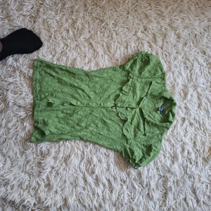 Grön spetsblus med volang och knappar - Söt grön blus från H&M med korta puffärmar, volangdetaljer vid kragen och knappar framtill. Blusen är i spetsliknande tyg med blommigt mönster och har en feminin, romantisk vibe. Perfekt för dig som gillar färg och detaljer.