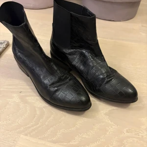 Svarta boots - Snygga svarta chelsea boots i crocopräglat skinn med låg klack och rund tå. Elastiska partier på sidorna gör dem enkla att ta på. Är i mycket bra skick