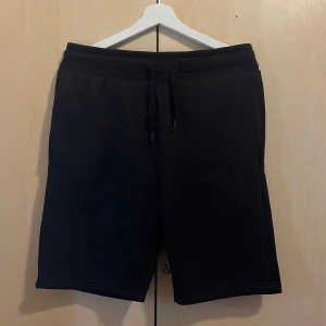 Svarta sweatshorts med snörning - Svarta shorts i mjuk bomull med relaxed fit och elastisk midja. Klassisk design med snörning i midjan och sidofickor. Perfekta för chill dagar eller träning. Tidlös look som funkar till det mesta.