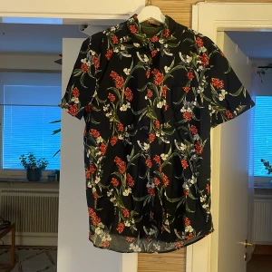Blommig svart skjorta Dressmann L - Svart kortärmad skjorta från Dressmann i slim fit-modell. Skjortan har ett färgstarkt blommönster i rött, vitt och grönt samt klassisk krage och knappar framtill. Perfekt för dig som vill sticka ut med en snygg och somrig vibe.