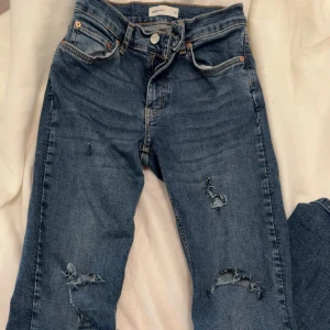Blå slitna jeans med hål - Säljer ett par blå jeans med snygga slitningar och hål på benen. Jeansen har klassisk femficksdesign, normal passform och är tillverkade i mjukt denimtyg. Dem är mid waist och har lite defekter 