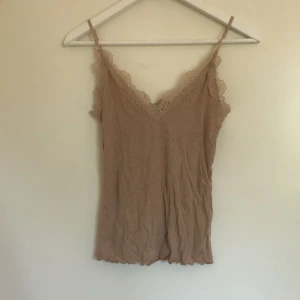 Beige ribbad linnetopp från new yorker❤️ - Supersöt beige linnetopp från new yorker med smala, justerbara axelband och v-ringning. Toppen är ribbad och har fina spetsdetaljer längs urringningen och axelbanden. Perfekt att styla med jeans eller kjol för en chill look.💐