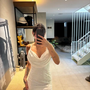 Vit pärlprydd kort klänning - Supersnygg vit kort klänning med pärldetaljer och draperad urringning. Klänningen har smala axelband och figurnära passform som framhäver siluetten. Perfekt för dig som vill sticka ut med en elegant och trendig look.