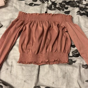 Rosa offshoulder topp med smock - Superfin offshoulder topp i dammigt rosa med långa ärmar och smockdetaljer upptill och nertill. Toppen har en lätt rynkad struktur och är perfekt för dig som gillar romantisk och trendig stil. Materialet känns mjukt och luftigt, och modellen är croppad med volangkant.