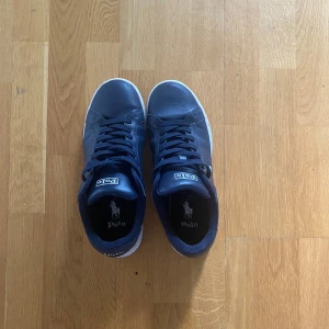Mörkblå sneakers från Polo Ralph Lauren - Snygga mörkblå sneakers från Polo Ralph Lauren med vit logga på sidan och vit sula. Skorna har klassisk rund tå, blåa snören och är tillverkade i skinn. Perfekta för en clean och stilren look.