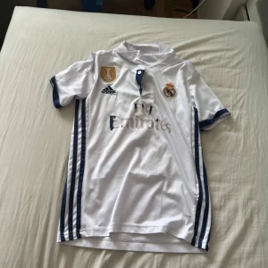 Real Madrid Ronaldo 7 Adidas tröja - Säljer en officiell Real Madrid fotbollströja från Adidas med Ronaldo 7 på ryggen. Tröjan är vit med mörkblå detaljer och ränder längs sidorna, broderad Real Madrid-text i nacken och klubbmärke på bröstet. Materialet är lätt och andas, perfekt för fotboll eller samling. Skadat tryck på rygg och bröst men annars bra kvalitet. 2016/17 hemma. Trycket kan återställas för 150kr extra