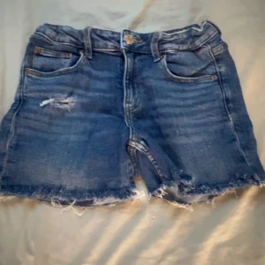 Blå jeansshorts  - Snygga blå jeansshorts med rå kant och slitningar framtill för en avslappnad vibe. Klassisk femficksmodell med knapp och dragkedja. Perfekta för varma dagar när du vill ha en cool och enkel stil.