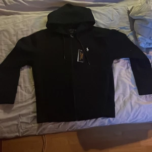 Svart hoodie från Polo Ralph Lauren - Svart hoodie från Polo Ralph Lauren med klassisk vit broderad logga på bröstet. Tröjan har huva med snörning och dragkedja framtill. Tillverkad i mjuk bomull, perfekt för en chill och stilren look, den är helt ny inte använt den nån gång 