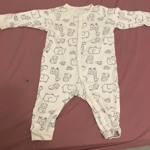 Vit byxdress med djurmönster - Supersöt vit byxdress med svarta tecknade djur som lejon, giraffer och noshörningar. Långärmad och helkropp med tryckknappar framtill för enkel påklädning. Mjuk och skön i bomull, perfekt för chill dagar hemma.