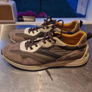 Bruna sneakers från Legend i mocka - Snygga sneakers från Legend i olika bruna nyanser med detaljer i beige och svart. Ovandelen är i mocka och skinn, med bruna snören och gul insida. Skorna har en sportig look med platt sula och rund tå, perfekta för en trendig vardagsstil. Använda 1 gång nya pris 1100